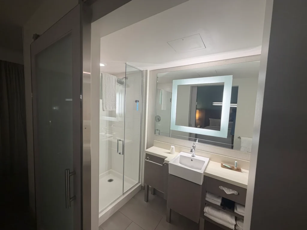 Bathroom at SpringHill Suites San Diego with a glass walk-in shower, square sink, lighted mirror, and open towel shelves